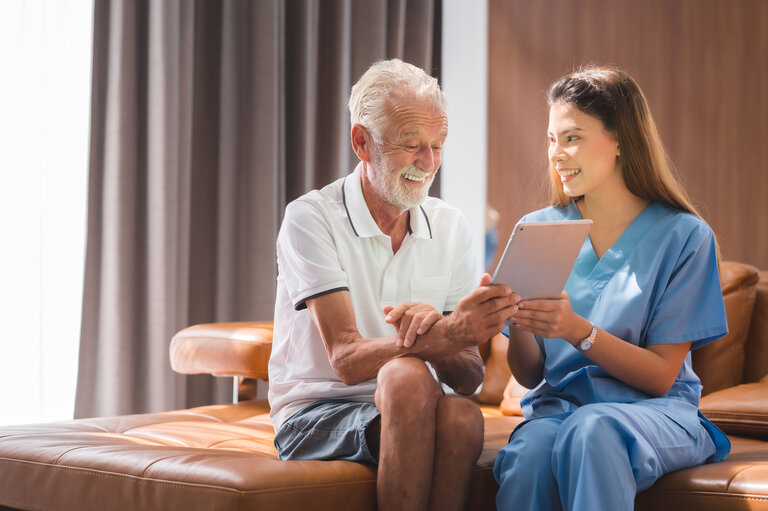 Krankenschwester oder Pflegerin, die einem älteren Mann den Umgang mit einem digitalen Tablet-Computer zu Hause beibringt