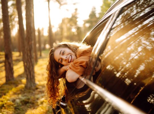 Junge Frau lächelt aus dem Autofenster, umgeben von Sonnenlicht und Waldlandschaft.