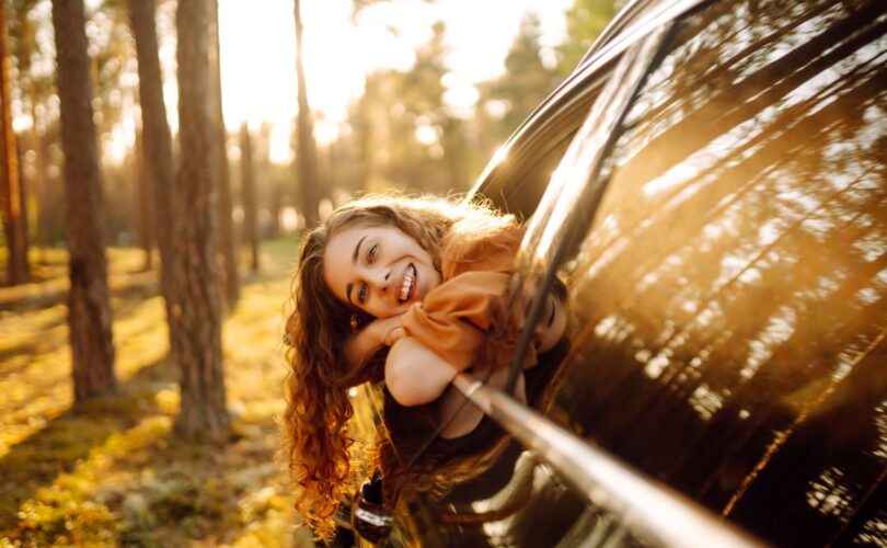 Junge Frau lächelt aus dem Autofenster, umgeben von Sonnenlicht und Waldlandschaft.