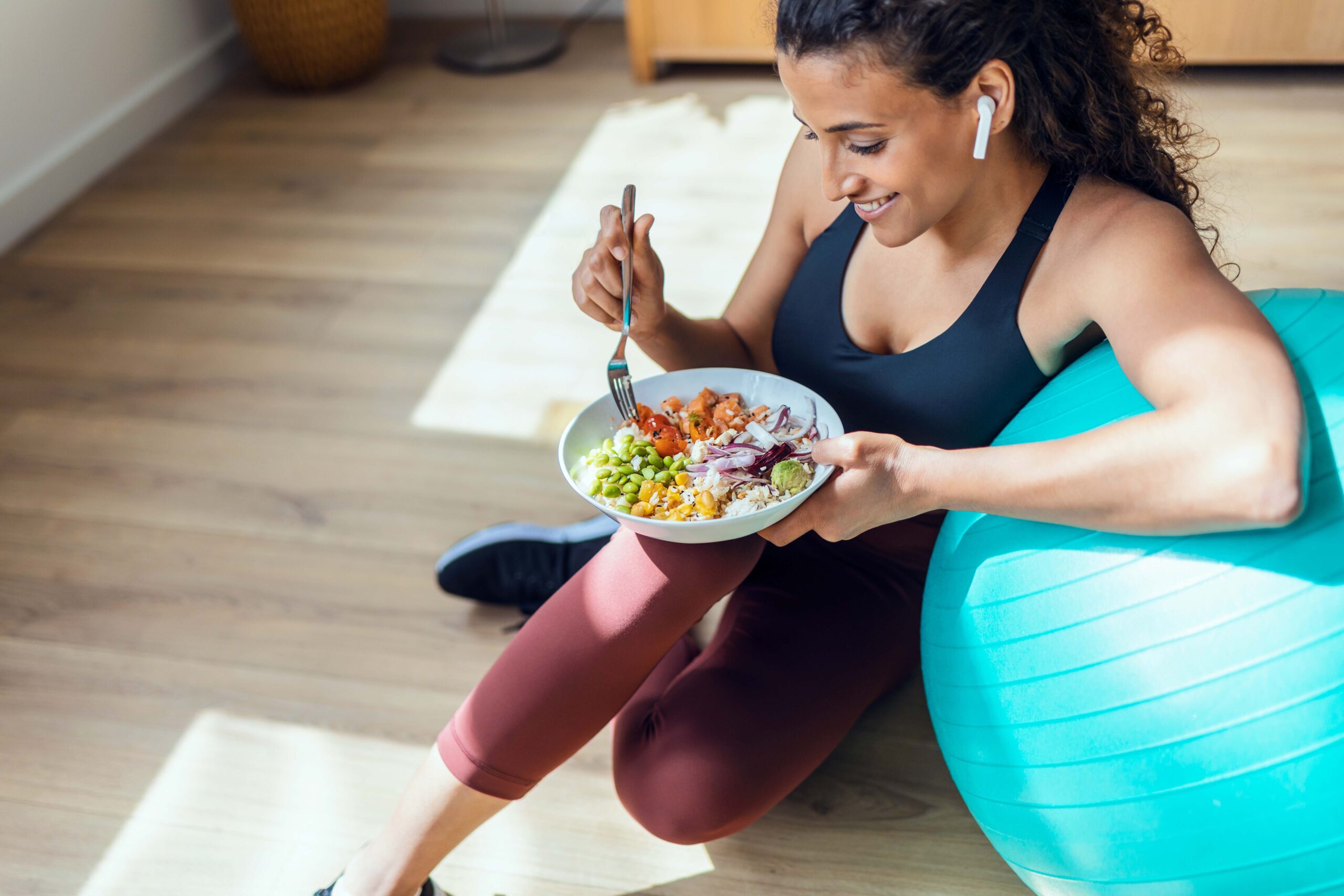 Sportliche junge Frau, die sich gesund ernährt, während sie zu Hause auf dem Boden sitzend Musik hört.