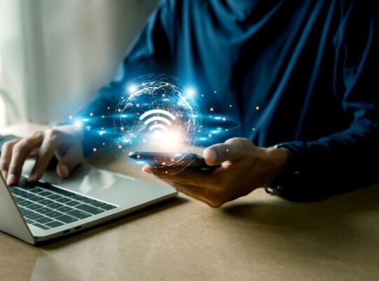 Person nutzt Laptop und Smartphone mit leuchtendem WLAN-Symbol – Symbolbild fuer MTP-basierte Netzwerktechnik und moderne Glasfaserinfrastruktur.
